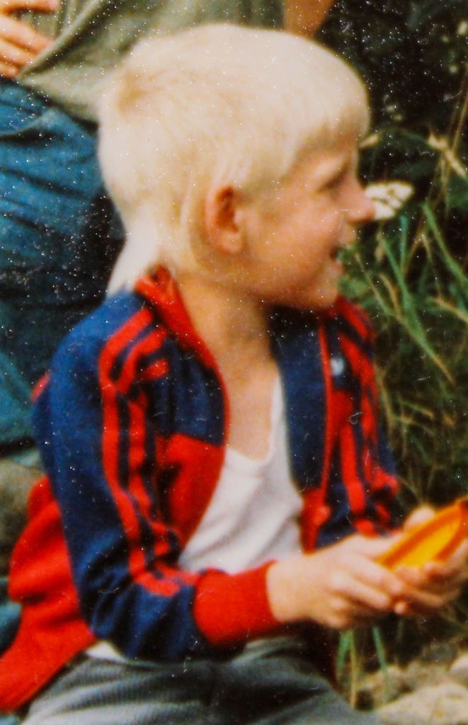 Nahaufnahme von mir als Grundschulkind auf dem Steg vor einem Teich hockend. Die Haare hellblond.