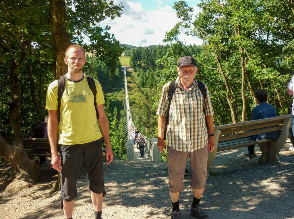 Links steh ich in gelbem T-Shirt und kurzer Hose, rechts mein Vater mit karriertem Hemd, Dreiviertelhose und Schirmmütze. Zwischen uns ist im Hintergrund eine lange Hängebrücke aus Stahl über einem grünen Tal zu sehen.