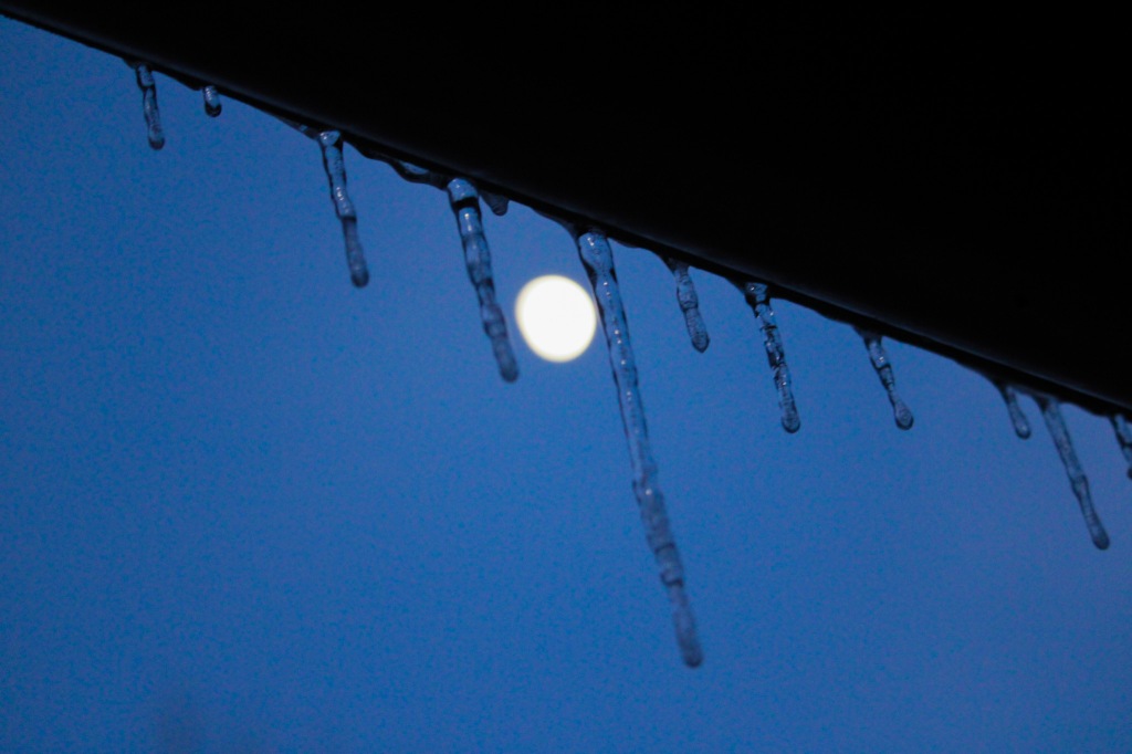 Eine Reihe von Eiszapfen an einer Regenrinne. Zwischen den beiden größten in der Mitte ist der Mond als kleiner Kreis vor einem dunkelblauen Abendhimmel zu sehen.
