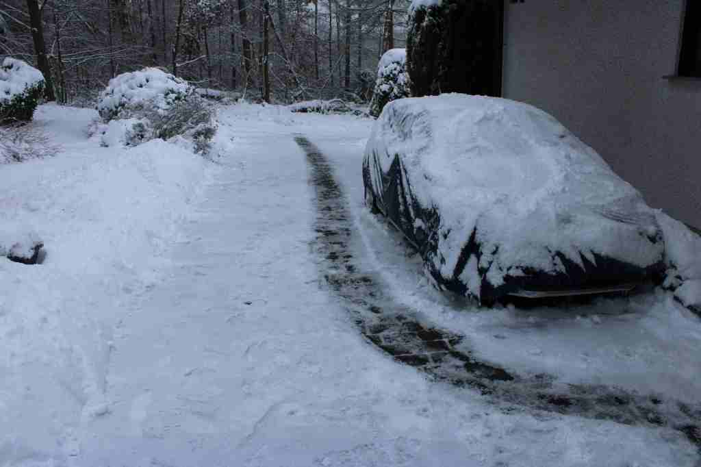 Die lange Hofeinfahrt. Rechts steht ein mit einer Plane abgedecktes und mit Schnee bedecktes Auto. Der Schnee im Hof ist weggeräumt, doch bis auf einen schmalen Laufweg in der MItte ist noch alles mit einer dicken Eissschicht bedeckt.