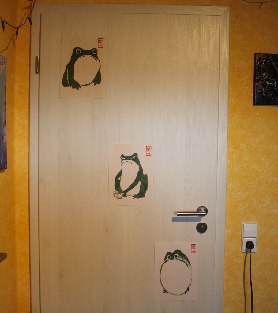 Eine weiße Zimmertür neben orangenfarbener Tapete. An der Tür befinden sich drei Drucke von gezeichneten Fröschen, die grimmig dreinblicken. Ein Bild oben links, eines in der Mitte und eines rechts unten unter dem Türgriff. Der Frosch in der Mitte bereitet einen Teezeremonie vor.