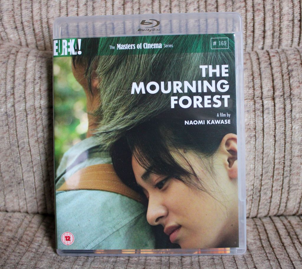 Blu-Ray-Box von "The Mourning Forst". Zu sehen ist eine Nahaufnahme der Hauptfigur Michiko, die von hinten den Kopf auf die Schulter des älteren Herrn legt, der wiederum nach oben in die Bäume schaut.