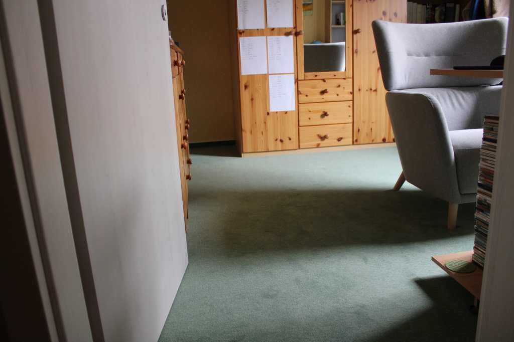 Blick aufs eingeräumte Arbeitszimmer mit dem neuen Teppichboden durch die Zimmertür. Rechts steht ein grauer Lesesessel, hinten an der Wand ein hellbraune Kleiderschrank.