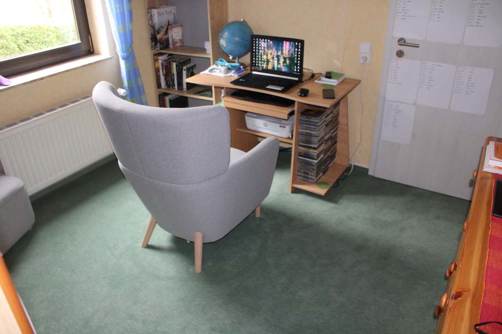 In der Mitte des Zimmers ein grauer Lesesessel von hinten, der vor einem kleinen Schreibtisch steht, auf dem ein Laptop aufgeklappt ist. Links steht ein Globus, noch weiter links davon ein Bücherregal, rechts die Zimmertür.