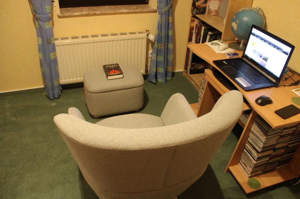 Der graue Lesesessel von hinten oben in Richtung Fenster und Heizung stehend, vor der ein passender grauer Fußhockersteht, auf dem ein Buch liegt. Rechts davon der Schreibtisch.