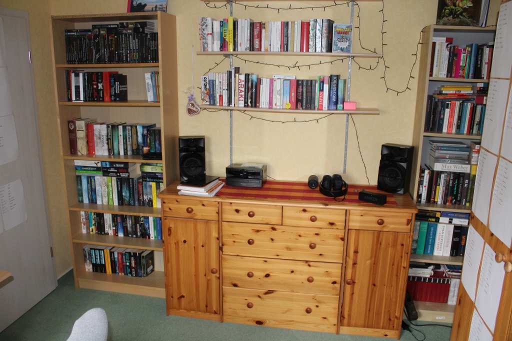 In der Mitte der Wand eine lange Kommodestehend, darauf eine kleine Stereoanlage. Darüber sind zwei Regealbretter mit Bücher zu sehen. Links und rechts davon zwei Billy-Bücherregale voll Bücher.