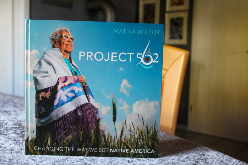 Foto vom aufrecht stehenden Buch "Project 562", darauf ist eine ältere indigene Frau mit modischer Brille, kurzen grauen Haaren und traditioneller Kleidung vor einem blauen Himmel in einem Weizenfeld zu sehen.