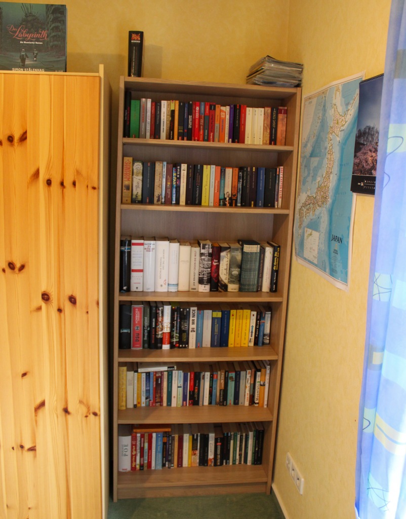 Ein Billy-Bücherrregal in der Zimmerecke. Rechtsdavon der Rand eines Kleiderschranks, links an der Wand hängt eine Landkarte von Japan.