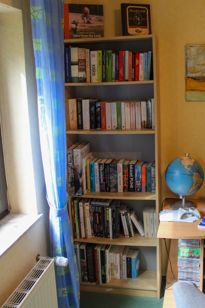 Ein Billy-Bücherrregal in einer Zimmerecke. Links ist noch das Fenster mit einem Vorhang und der Heizung zu sehen. Rechts der Rand eines Schreibtischs auf dem ein Globus steht.